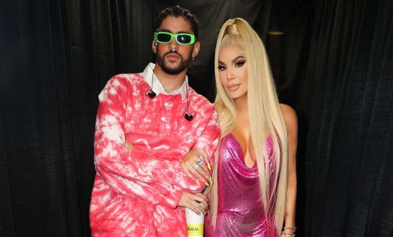 A man and a woman posing together, the man in a pink tie-dye hoodie and sunglasses, and the woman in a metallic pink dress with long blonde hair, standing in an area draped with black