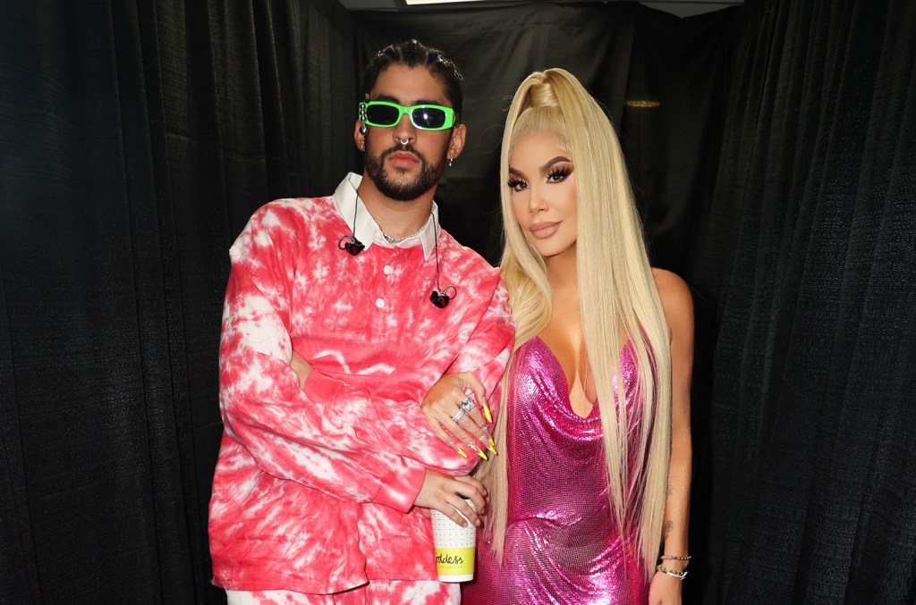 A man and a woman posing together, the man in a pink tie-dye hoodie and sunglasses, and the woman in a metallic pink dress with long blonde hair, standing in an area draped with black