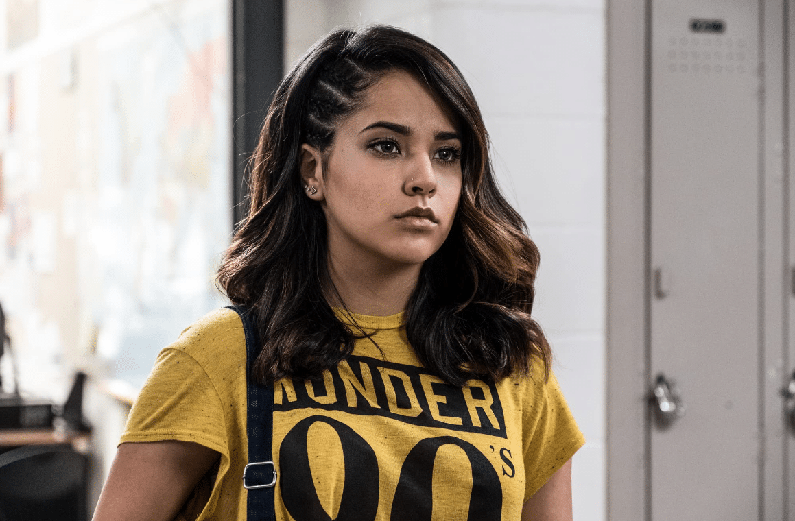 Becky G, with medium-length dark hair styled in braids, looks intensely to the side. She wears a vibrant yellow t-shirt with "thunder" written in black, standing in a