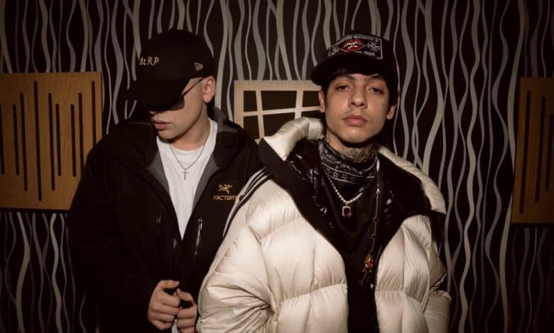 Two young men standing against a patterned backdrop; one in a white puffer jacket and black scarf, the other in a black jacket and cap, both looking cool and confident during the Bizarrap