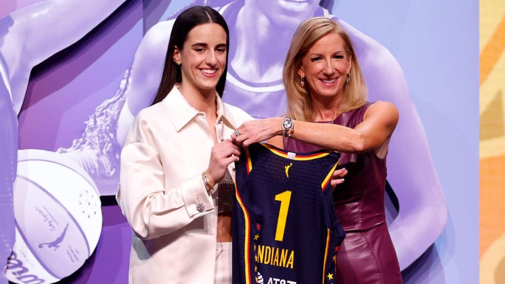 Two women, smiling, stand side by side at a sports event. One is presenting the other with a record-breaking basketball jersey that features "Indiana" and a bold number 1. A promotional basketball