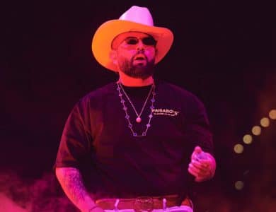 A man performs on stage, wearing a white cowboy hat, black shirt with "paisaboys" logo, silver necklace, and belt. red lighting bathes the scene, emphasizing his dynamic presence.