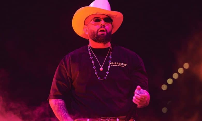 A man performs on stage, wearing a white cowboy hat, black shirt with "paisaboys" logo, silver necklace, and belt. red lighting bathes the scene, emphasizing his dynamic presence.