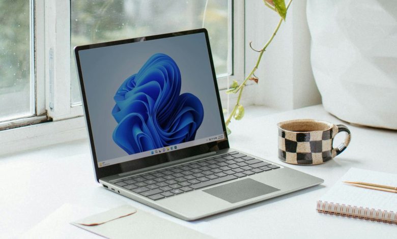 A laptop with a blue abstract wallpaper on its screen, placed on a desk near a window displaying "Easy Methods to Enhance Windows Security." Next to it are a polka dot mug, a house