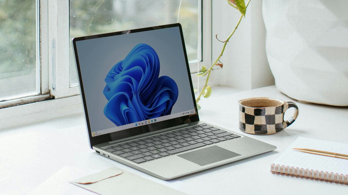 A laptop with a blue abstract wallpaper on its screen, placed on a desk near a window displaying "Easy Methods to Enhance Windows Security." Next to it are a polka dot mug, a house