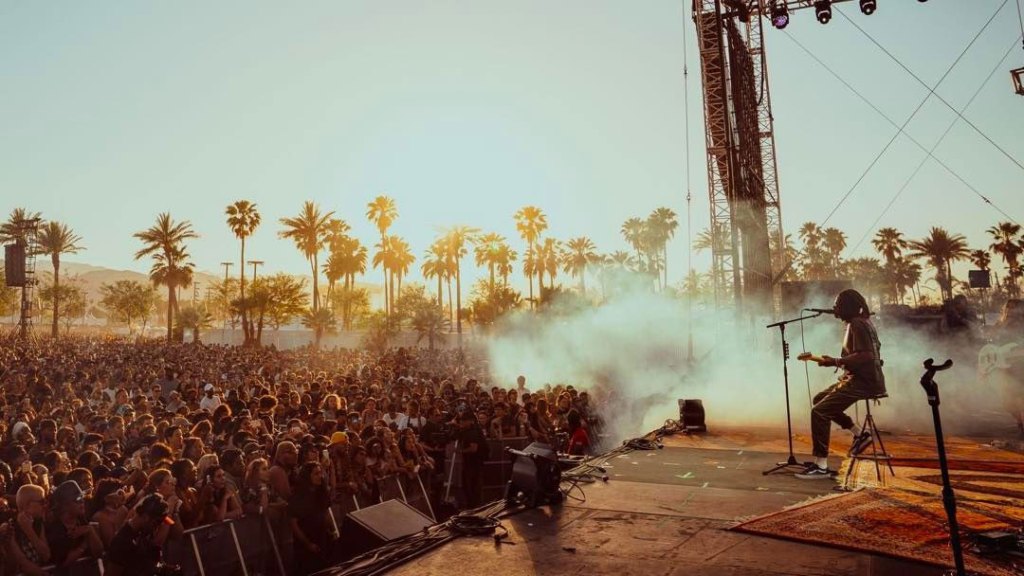 A musician with long hair plays an electric guitar on stage at a top 2024 music festival during sunset, with a large crowd of spectators and palm trees in the background.