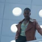 A man standing on a balcony with a modern design, featuring large circular lights on the ceiling. He wears a brown jacket, striped shirt, and has a confident expression befitting Doctor Who. The