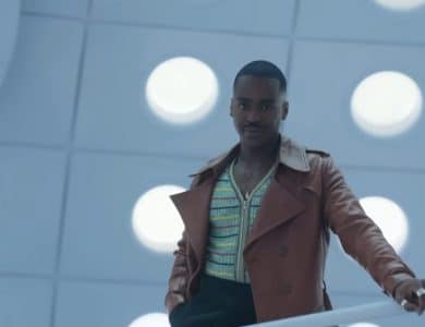 A man standing on a balcony with a modern design, featuring large circular lights on the ceiling. He wears a brown jacket, striped shirt, and has a confident expression befitting Doctor Who. The