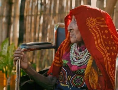An elderly woman in a wheelchair, wearing a vibrant red and gold traditional outfit and heavy silver jewelry, smiles gently while holding a cane, with a background of sunlit bamboo at the Panama Film Fest.