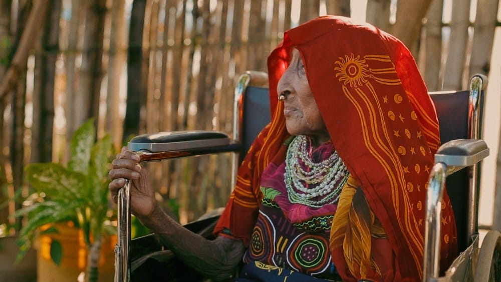 An elderly woman in a wheelchair, wearing a vibrant red and gold traditional outfit and heavy silver jewelry, smiles gently while holding a cane, with a background of sunlit bamboo at the Panama Film Fest.