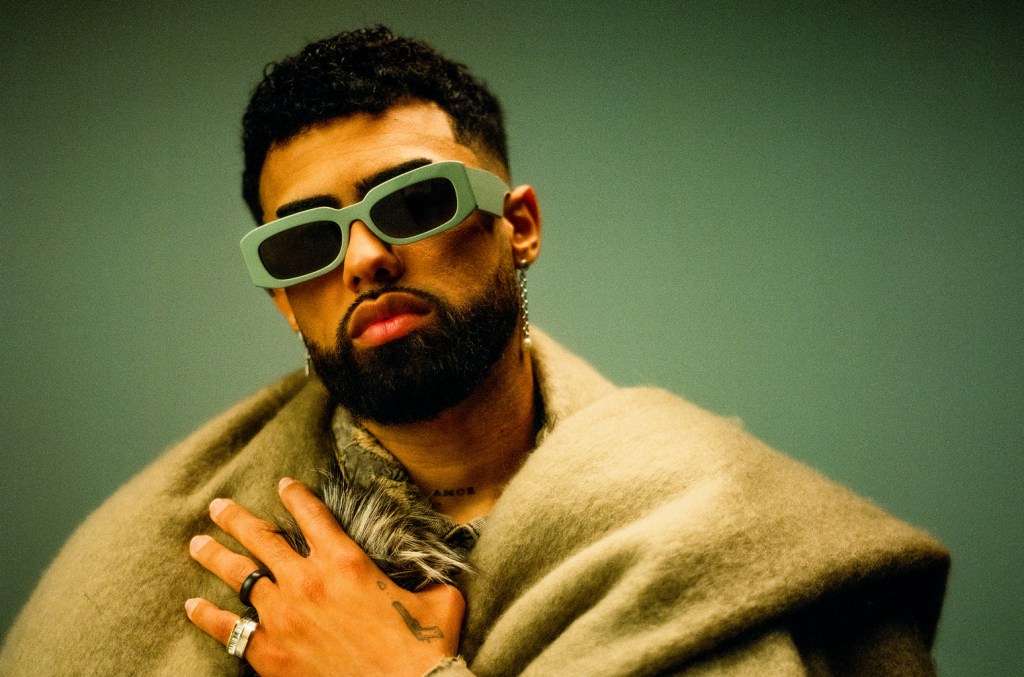 A stylish man with a beard poses confidently, wearing oversized gray sunglasses and a luxurious beige shawl over a fur coat. His hand, adorned with a large ring, rests on his chest, displaying a