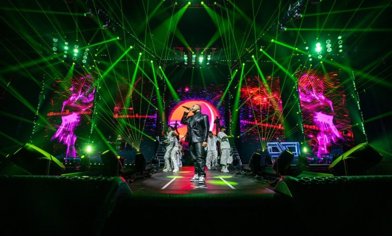A dynamic concert scene with Don Omar in a black and white outfit and cap standing center stage, backlit by a vibrant display of green laser beams and illuminated geometric patterns. Two large screens show silhou