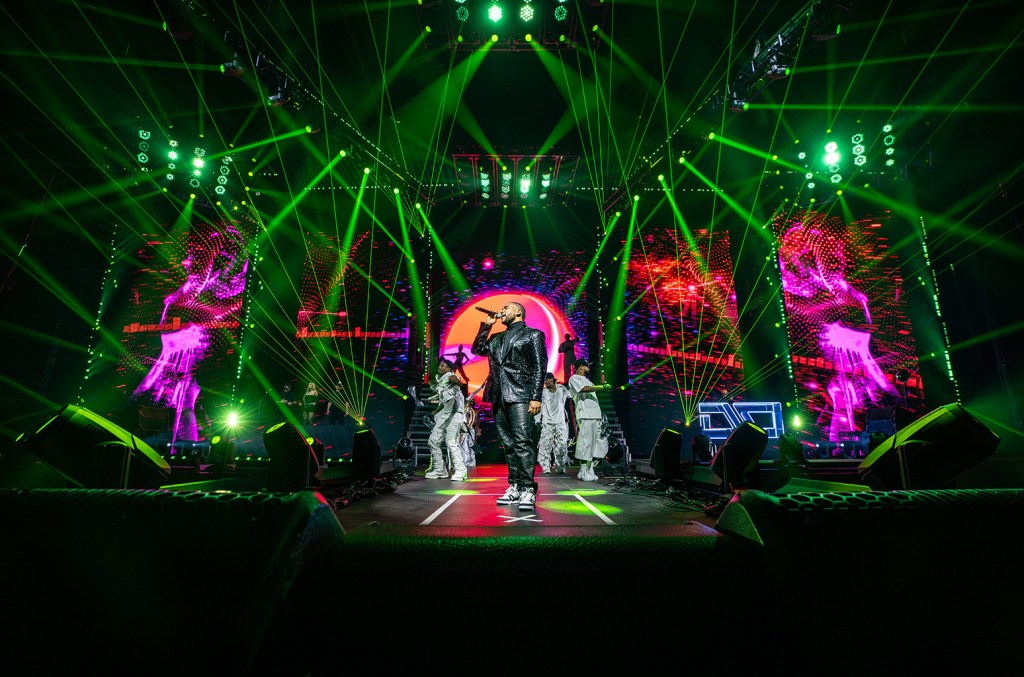 A dynamic concert scene with Don Omar in a black and white outfit and cap standing center stage, backlit by a vibrant display of green laser beams and illuminated geometric patterns. Two large screens show silhou