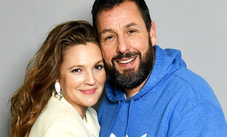 A smiling man in a blue hoodie embraces Drew Barrymore, who is in a cream blazer. They appear cheerful and affectionate, posing closely in a studio setting with a gray background.