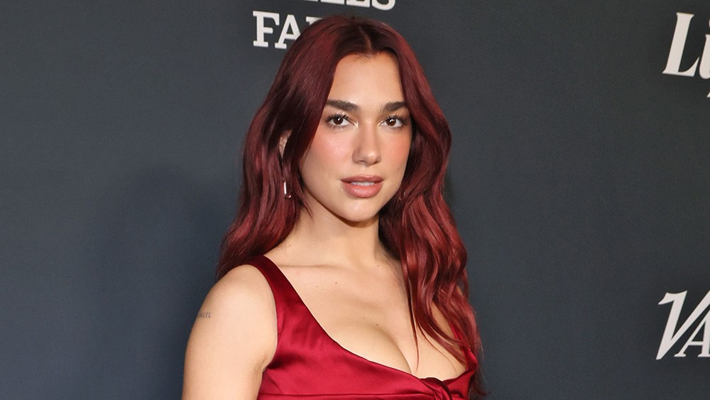 A woman with long, wavy red hair and wearing a deep red satin dress poses at an event hosted by Dua Lipa, standing against a dark background with visible logos. She has a subtle
