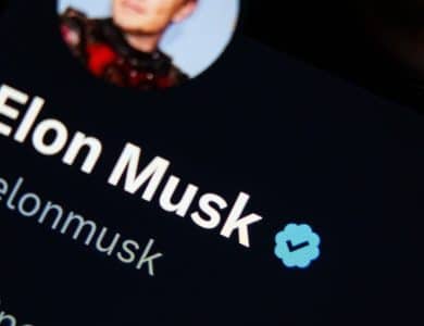 Close-up of a digital screen displaying Elon Musk's Verified X profile with his username @elonmusk and a part of his profile picture showing him in a red outfit.