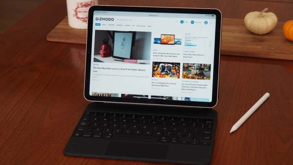 A tablet with a keyboard attachment on a wooden table, displaying the Gizmodo website. A stylus and a small pumpkin are also on the table, suggesting a cozy, autumnal setting.