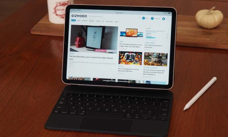 A tablet with a keyboard attachment on a wooden table, displaying the Gizmodo website. A stylus and a small pumpkin are also on the table, suggesting a cozy, autumnal setting.