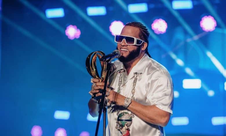 A man stands on stage holding a trophy at the 10th Anniversary Celebration of Heat Latin Music Awards, wearing a white shirt, multiple necklaces, and blue sunglasses under vibrant purple stage lights. His