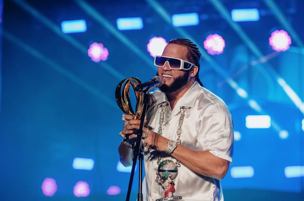 A man stands on stage holding a trophy at the 10th Anniversary Celebration of Heat Latin Music Awards, wearing a white shirt, multiple necklaces, and blue sunglasses under vibrant purple stage lights. His