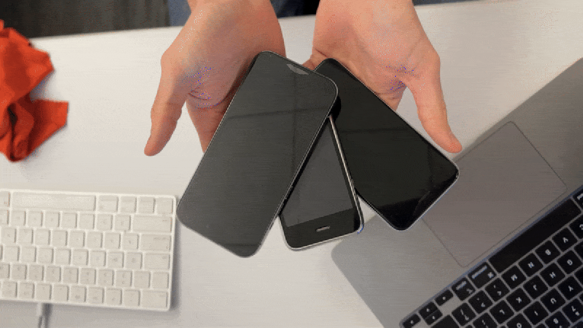 A person's hands are positioned above a desk, about to choose between three smartphones. The phones are black, lying face down. A laptop and A Handbook for the Enlighteningly Uninformed