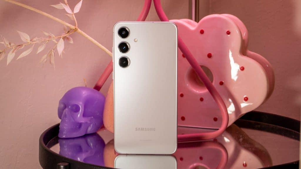 A Samsung smartphone with a white back and triple camera setup rests on a glass surface, surrounded by a pink neon heart light, purple skull, and pink cherry blossoms, illustrating the step-by-step guide