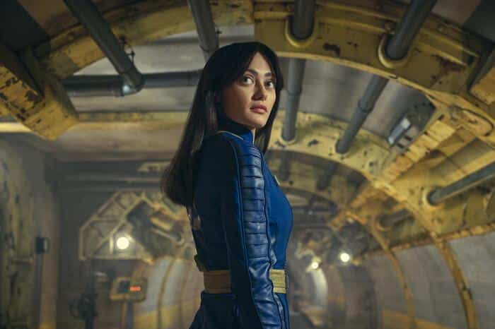 A woman in a blue suit stands confidently inside a yellow, industrial-looking tunnel with pipes and metallic textures. her expression is serious, and she has long dark hair. soft lighting highlights her from the side.