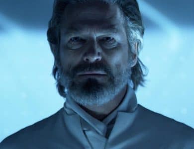 A middle-aged man with a full beard and mustache looks intently forward. he wears a high-collared grey outfit. the lighting casts a blue hue over the scene, emphasizing the seriousness of his expression.