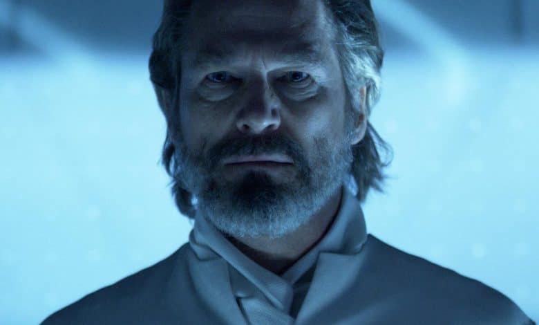 A middle-aged man with a full beard and mustache looks intently forward. he wears a high-collared grey outfit. the lighting casts a blue hue over the scene, emphasizing the seriousness of his expression.