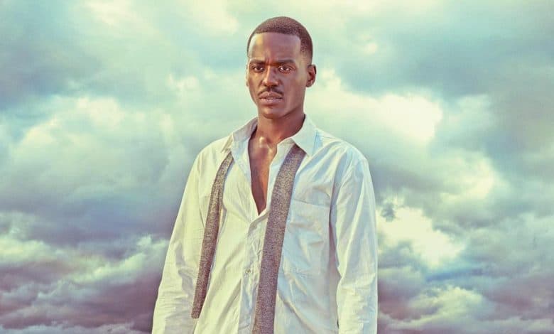 A young man stands against a dramatic cloudy sky. he wears an unbuttoned white shirt over a light grey tee, gazing directly at the camera with a serious expression. the lighting accentuates his facial features.