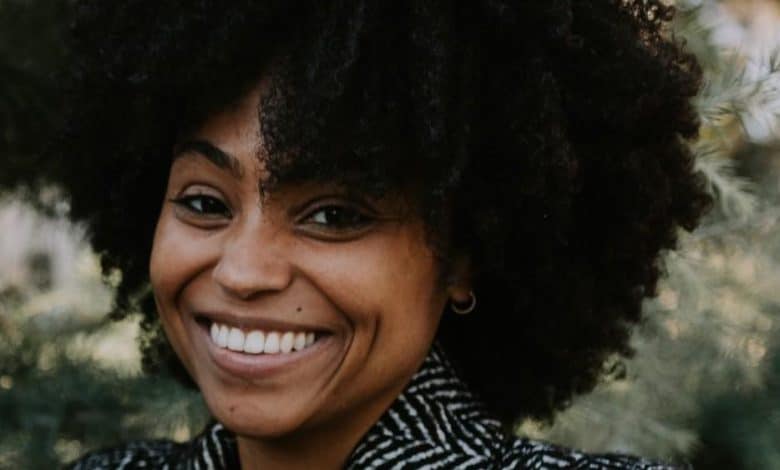 A cheerful black woman with a broad smile, sporting a large, curly afro. she wears a striped jacket and stands against a blurred greenery background.