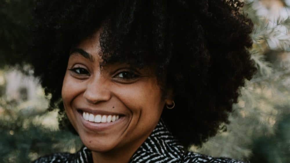 A cheerful black woman with a broad smile, sporting a large, curly afro. she wears a striped jacket and stands against a blurred greenery background.