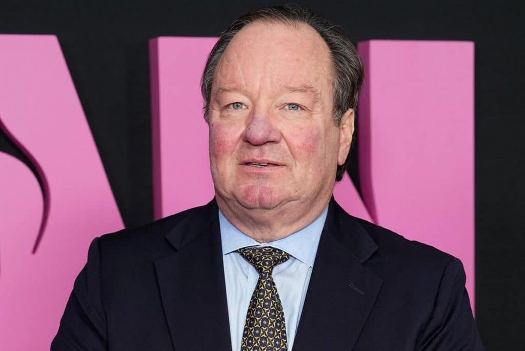 A middle-aged man in a dark suit, white shirt, and patterned tie smiles at a pink-themed event, with a partially visible pink backdrop marked with large black letters.