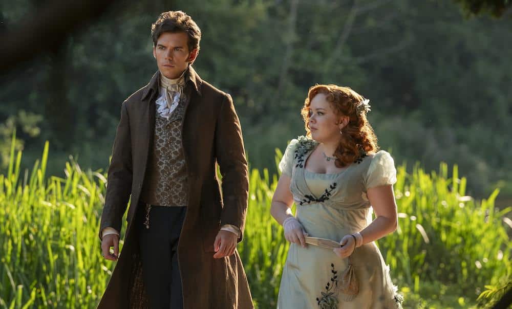 A man and a woman in early 19th-century attire walk through a sunlit field. the man wears a dark coat and vest, while the woman, holding a book, wears a light blue empire-waist dress with floral details.
