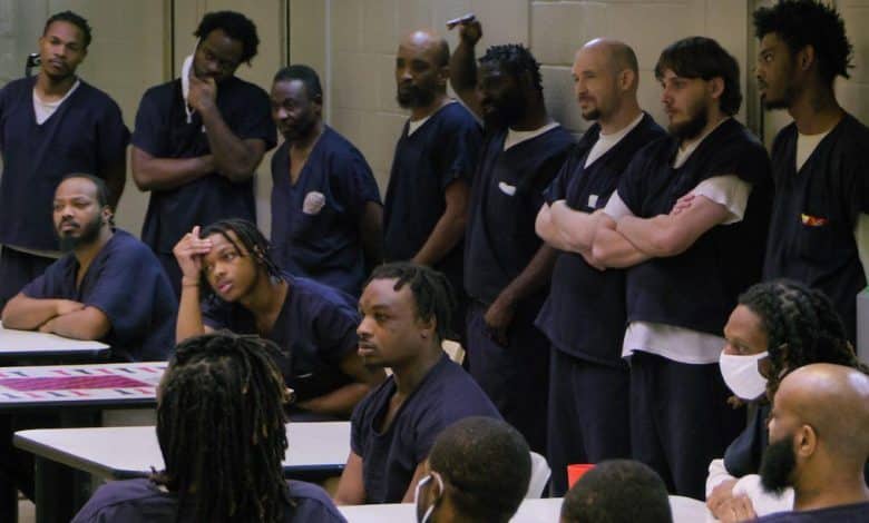 A group of incarcerated men in blue uniforms gathers around tables in a room, listening intently to a speaker out of frame. some stand, while others sit or lean on tables, showing various expressions of engagement and contemplation.