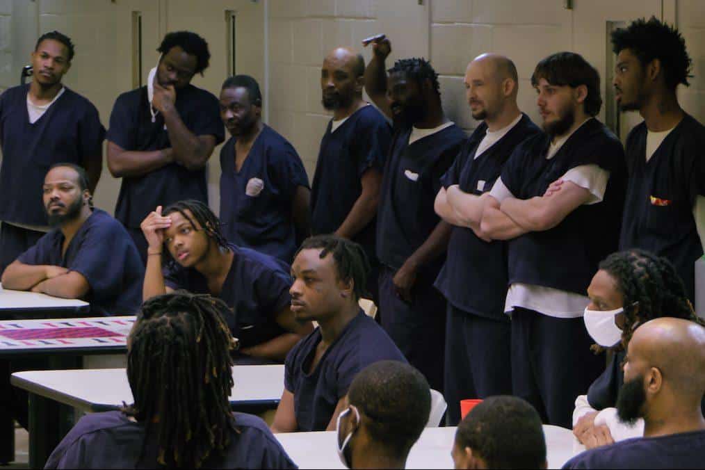 A group of incarcerated men in blue uniforms gathers around tables in a room, listening intently to a speaker out of frame. some stand, while others sit or lean on tables, showing various expressions of engagement and contemplation.