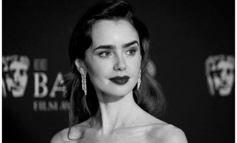 A black and white portrait of a woman with dark, slicked-back hair and bold lipstick at a formal event, wearing a glamorous earring and gazing to the side against a backdrop featuring logos.