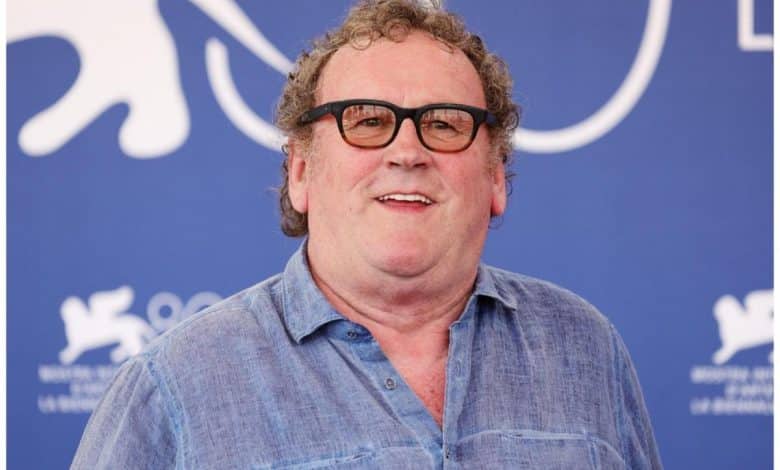 A middle-aged man with curly hair and glasses smiling at a blue event backdrop. he wears a light blue, button-down shirt.