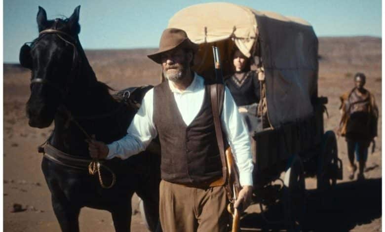 A man in early 20th-century attire, including a hat and vest, leads a black horse pulling a covered wagon across a dusty terrain. two people are visible inside the wagon, and another person follows on foot in the background.