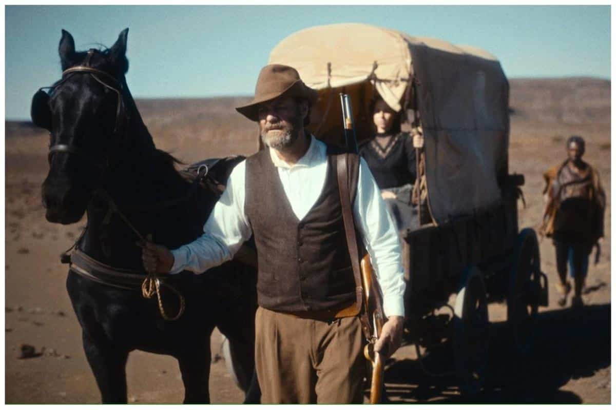 A man in early 20th-century attire, including a hat and vest, leads a black horse pulling a covered wagon across a dusty terrain. two people are visible inside the wagon, and another person follows on foot in the background.