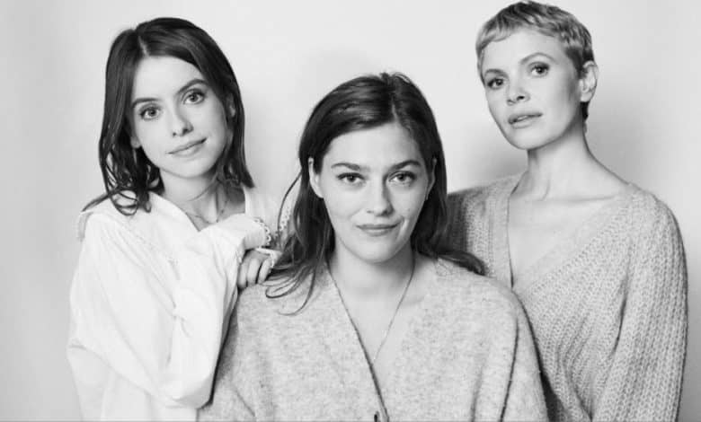 Three women of varied appearances stand close together against a light background, smiling subtly at the camera. they wear casual, cozy clothes, ranging from a white blouse to soft sweaters.