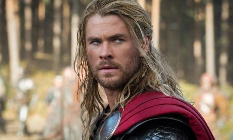 A close-up of a man with long blond hair and a beard, wearing medieval armor and a red cape, stands solemnly in a forest setting, looking intently towards the camera.