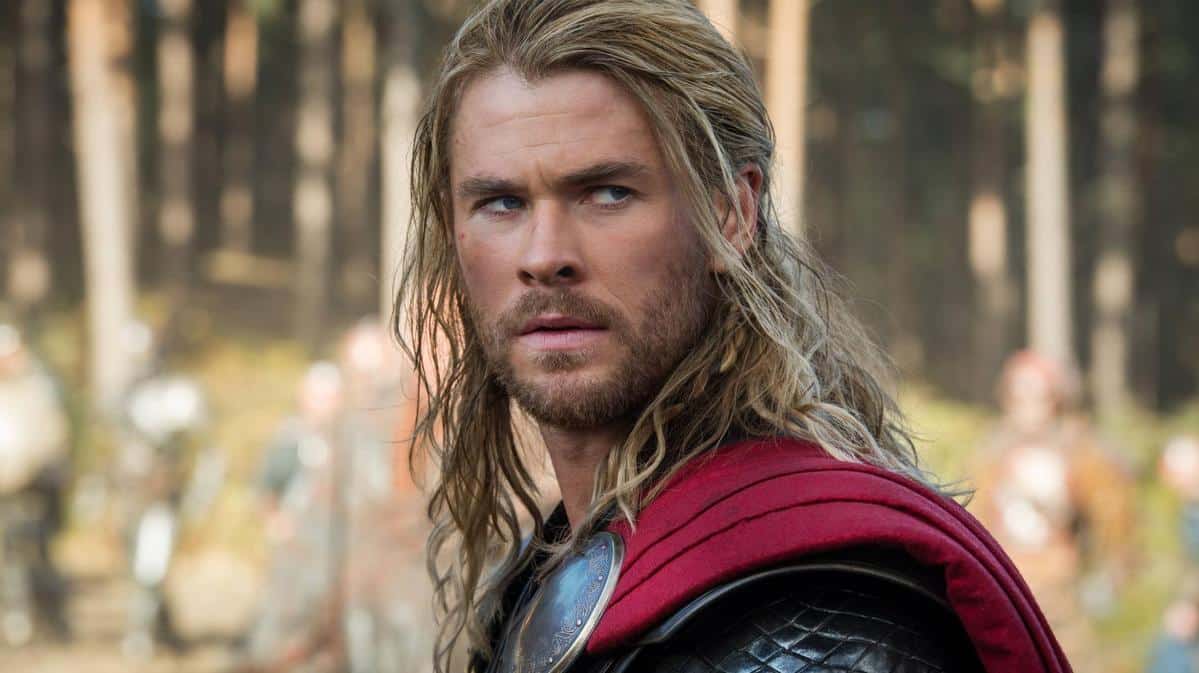 A close-up of a man with long blond hair and a beard, wearing medieval armor and a red cape, stands solemnly in a forest setting, looking intently towards the camera.