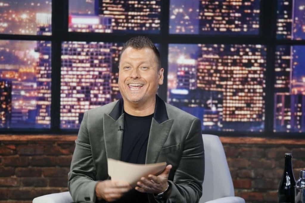 A man with short hair sits smiling in a tv studio. he's wearing a dark blazer over a gray shirt, holding papers, with a nighttime cityscape backdrop lit with many lights.