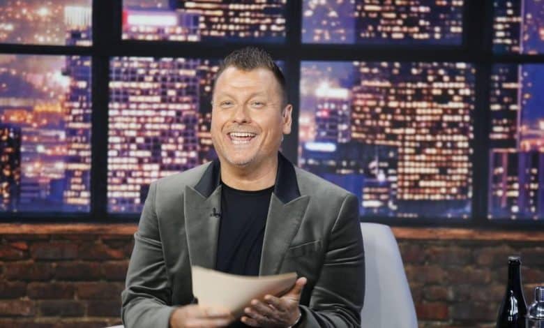 A man with short hair sits smiling in a tv studio. he's wearing a dark blazer over a gray shirt, holding papers, with a nighttime cityscape backdrop lit with many lights.