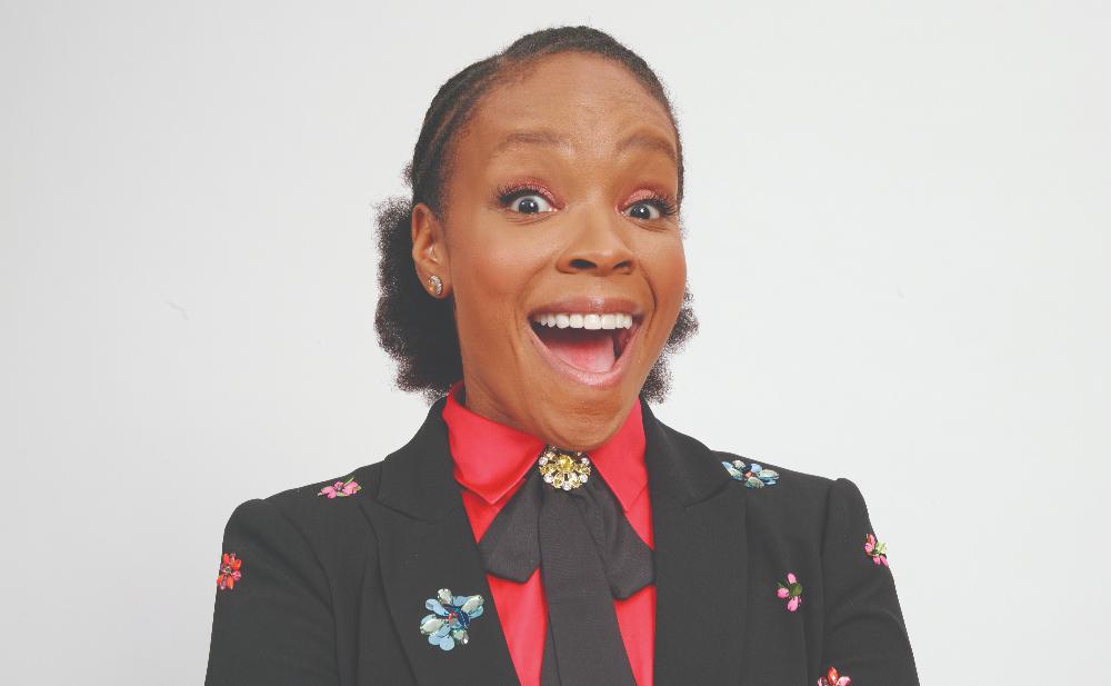A joyful african american woman with her mouth wide open in excitement, wearing a black blazer with colorful floral embroidery and a red blouse, against a white background.