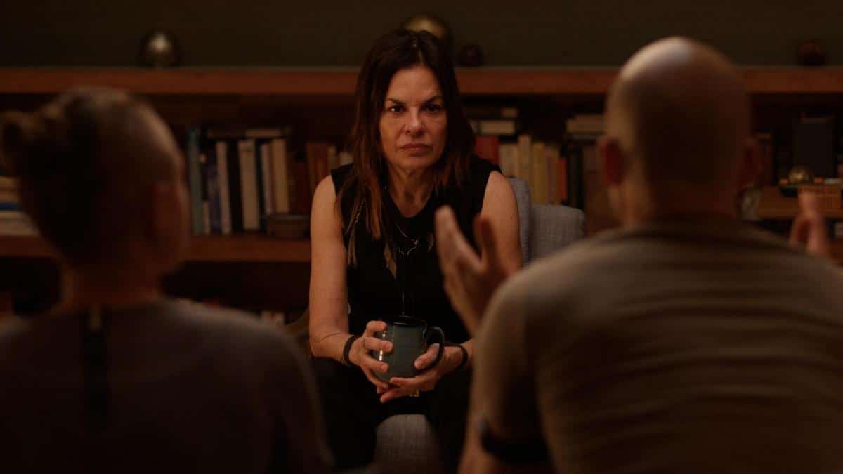 A woman with shoulder-length brown hair sits holding a mug, engrossed in a group discussion in a cozy, dimly lit room filled with books. two blurred figures face her, engaging her attention, highlighting an intimate, conversational atmosphere.