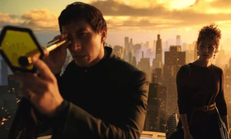 A man in a black suit examines a hexagonal device, while a woman with short, curly hair in a dark dress stands beside him, both overlooking a cityscape at sunset.