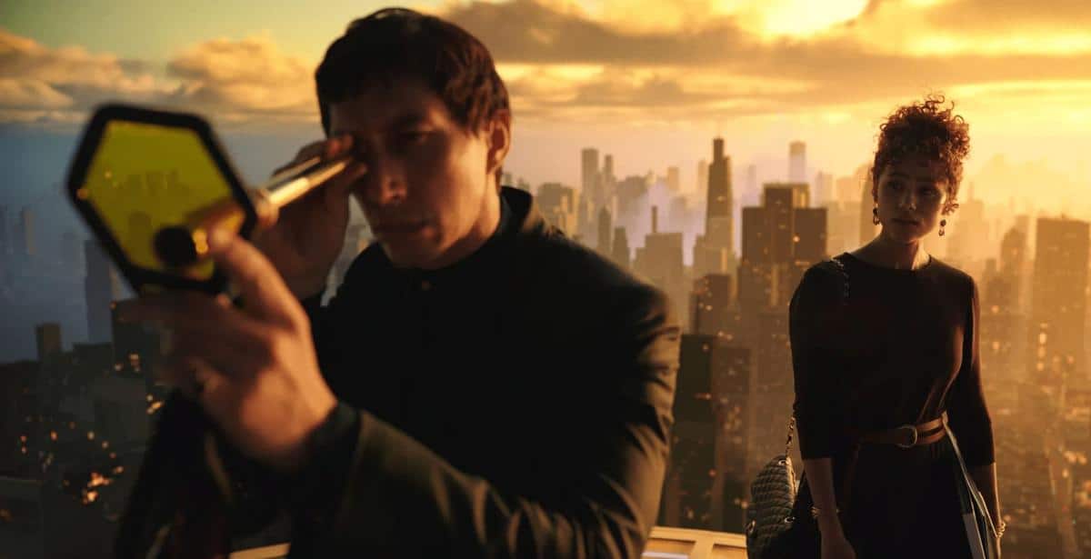 A man in a black suit examines a hexagonal device, while a woman with short, curly hair in a dark dress stands beside him, both overlooking a cityscape at sunset.