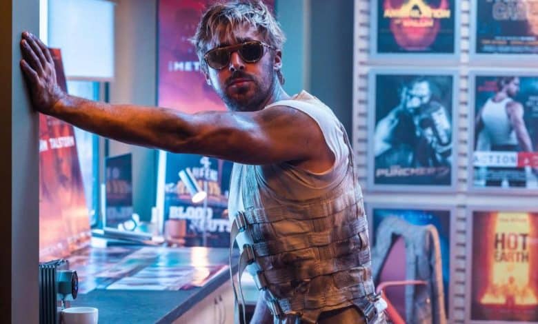 A man with tousled blond hair and sunglasses poses in a sleeveless top and tactical vest in a room filled with movie posters and neon signs, exuding a rugged, action-packed vibe.